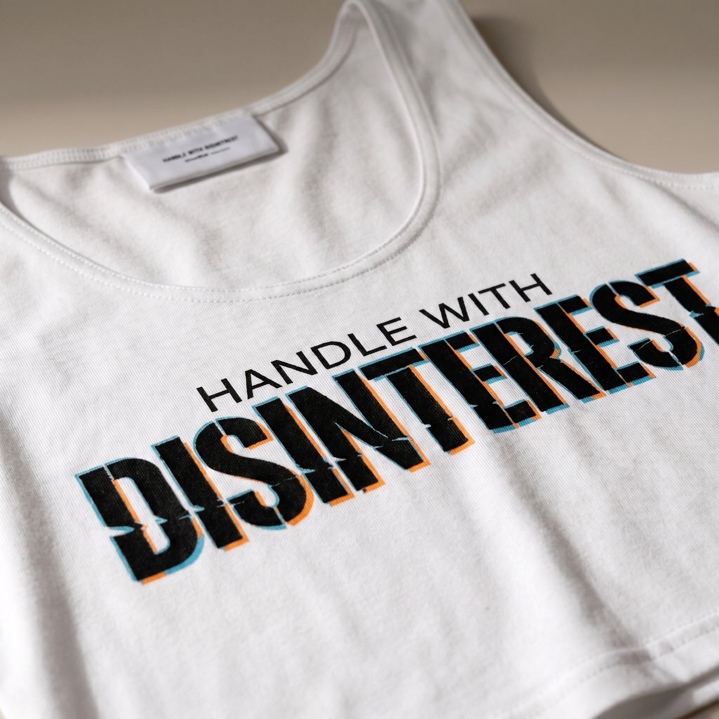 White tank top with 'HANDLE WITH DISINTEREST' text on a neutral background
