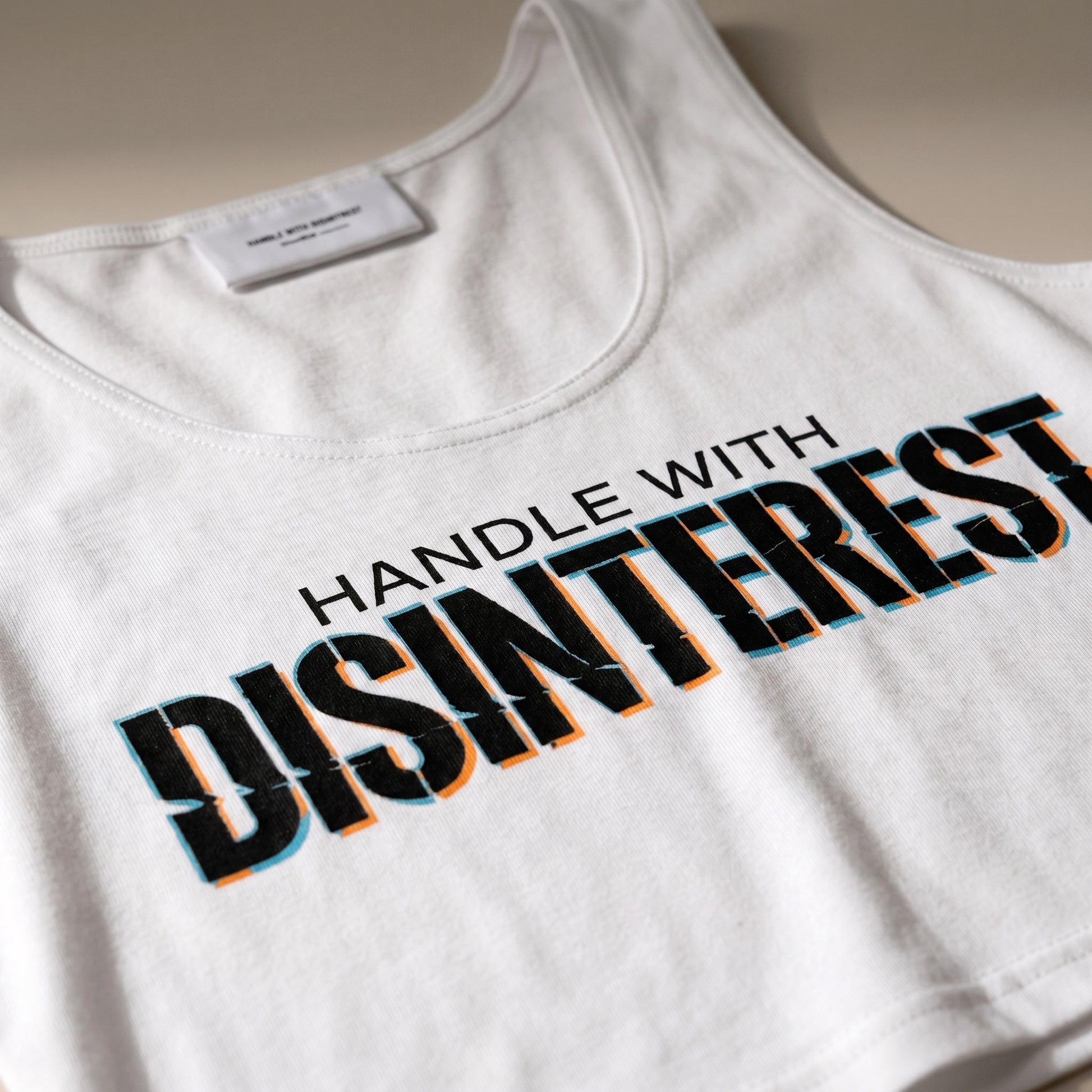 White tank top with 'HANDLE WITH DISINTEREST' text on a neutral background