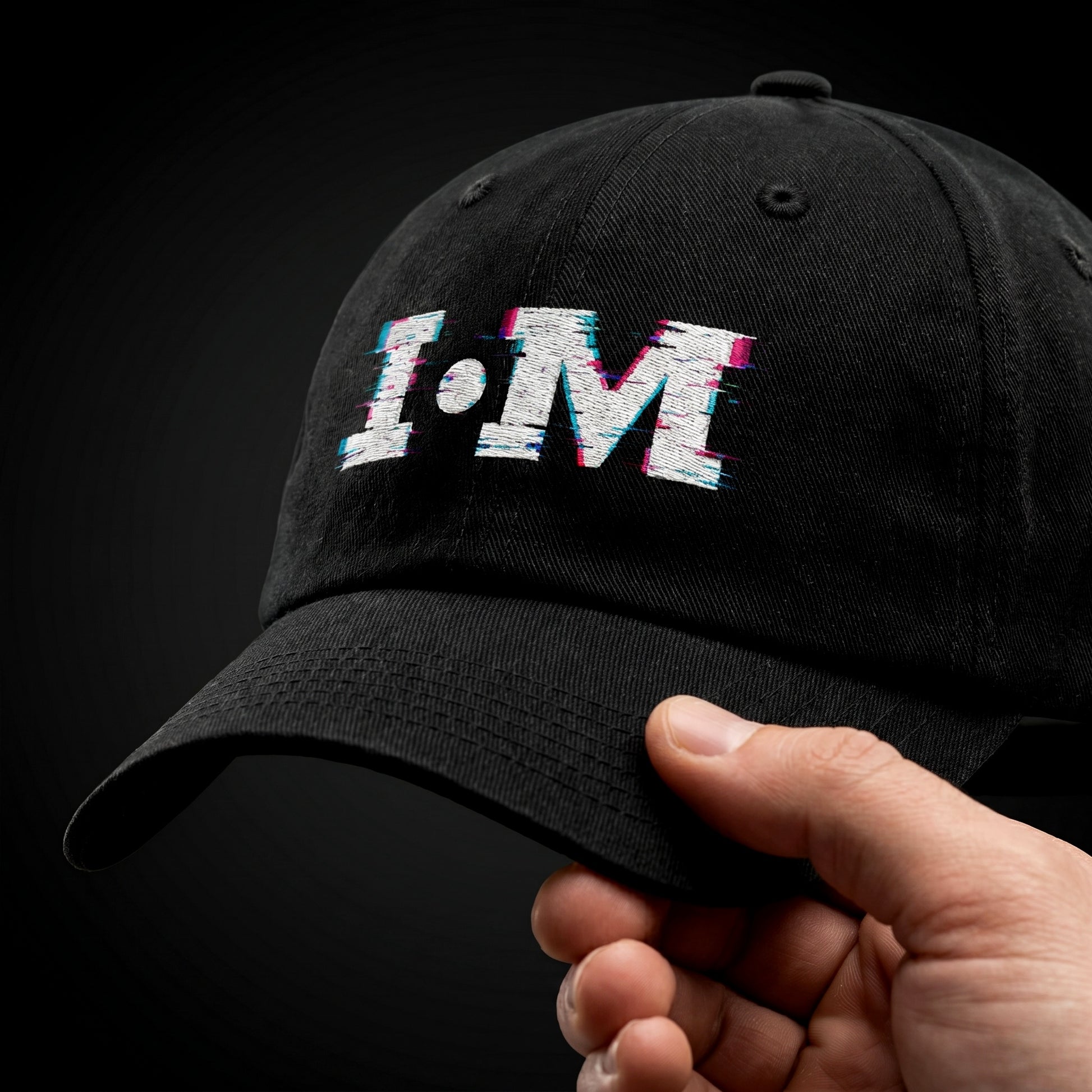 Black cap with 'I·M' text held by a hand against a dark background