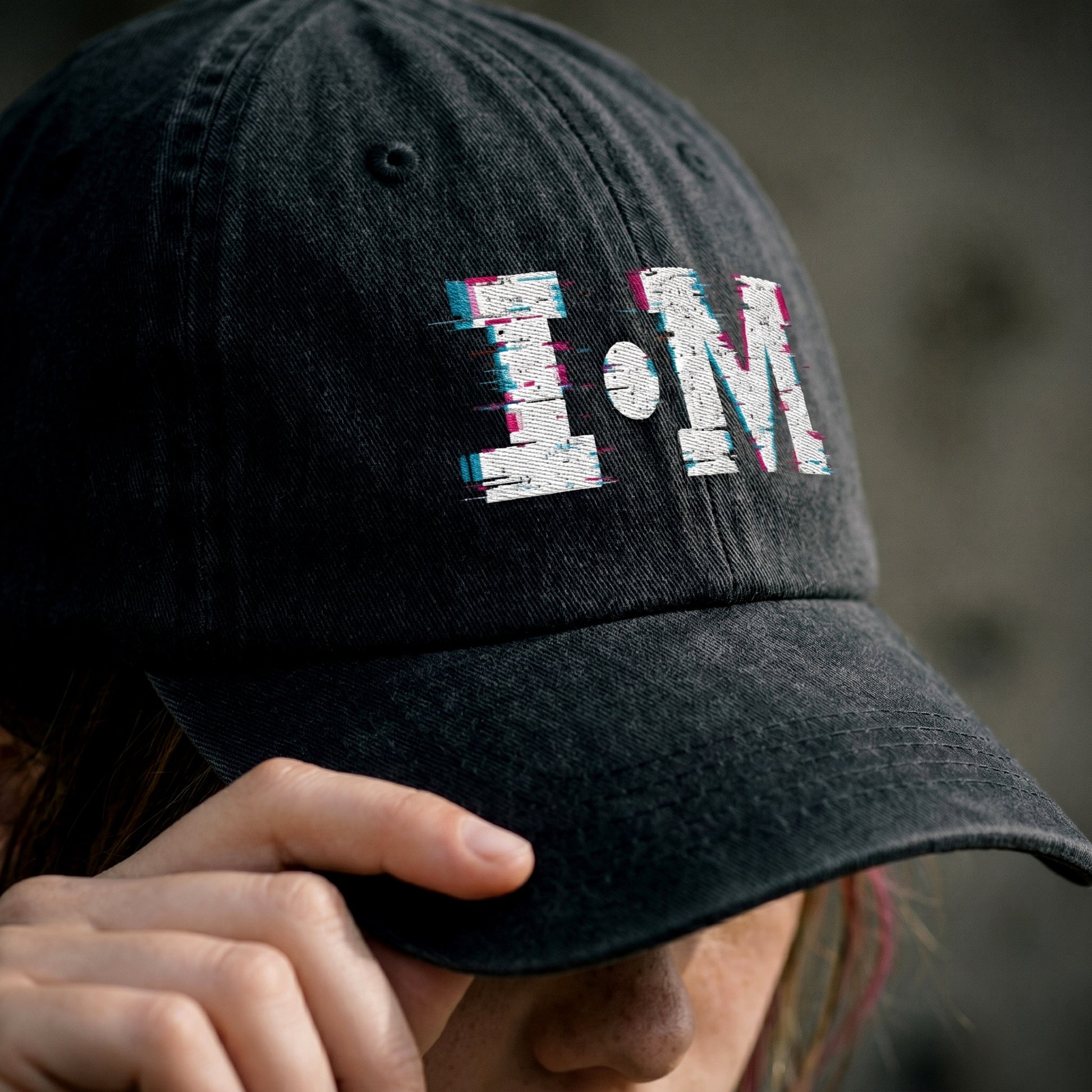 Black cap with 'I.M' logo held by a person against a blurred background