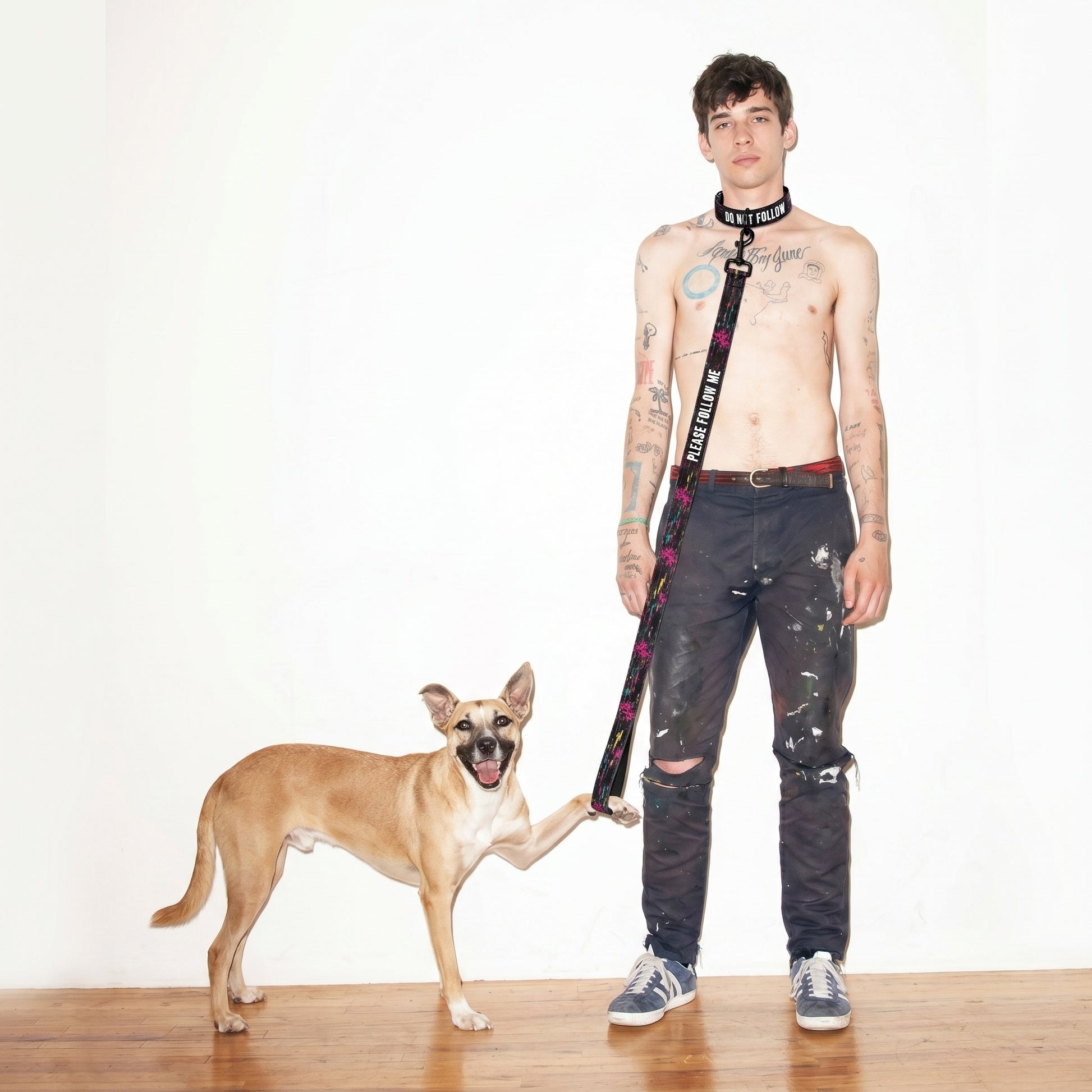 Man with tattoos and a dog standing on a wooden floor against a white wall