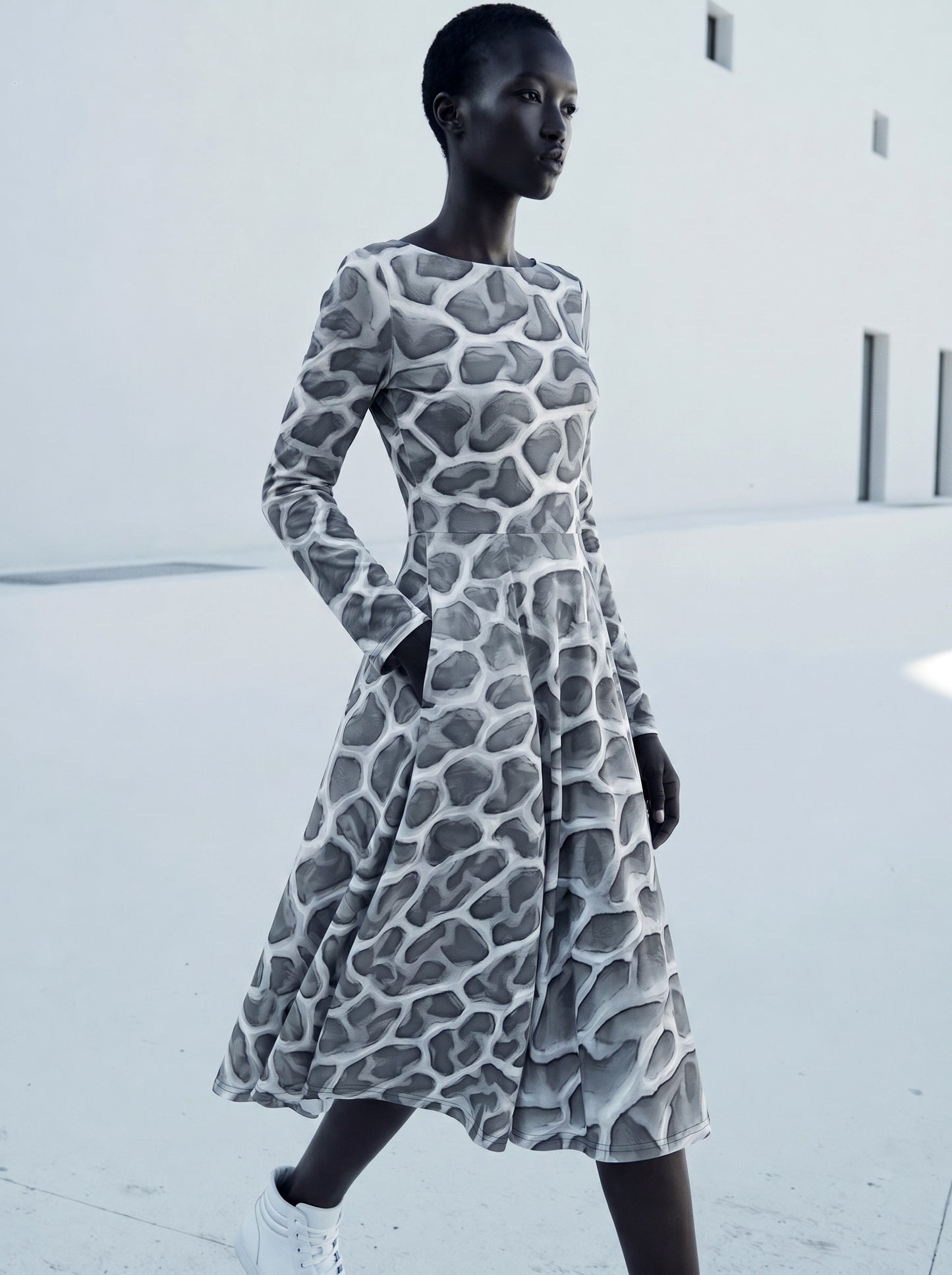 Editorial fashion image of a model wearing the ANSO Cellular Mesh Midi Dress against a warm neutral studio background, showing the long sleeves, flared midi skirt, and sculptural grey mesh print.