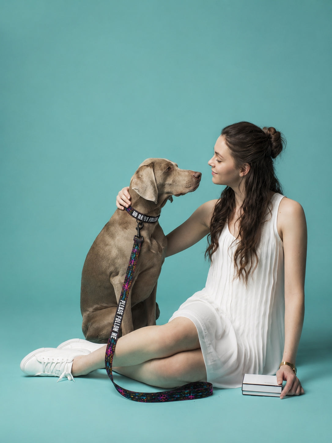 Woman in a white dress sitting on the ground with a brown dog against a teal background