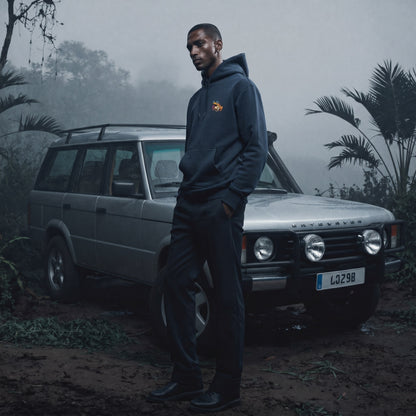 Man in a dark hoodie standing next to a Land Rover in a misty forest setting