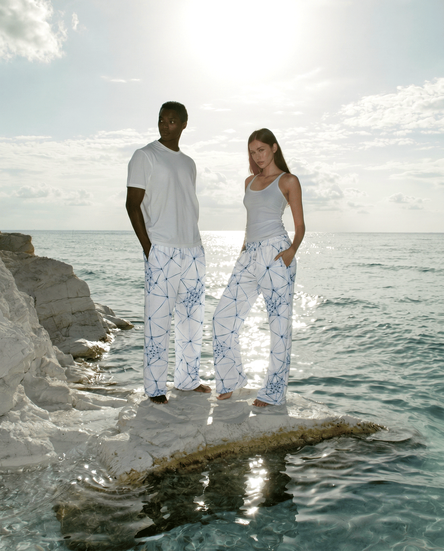 Two people standing on a rock by the ocean wearing white ANSO joggers with blue patterns.