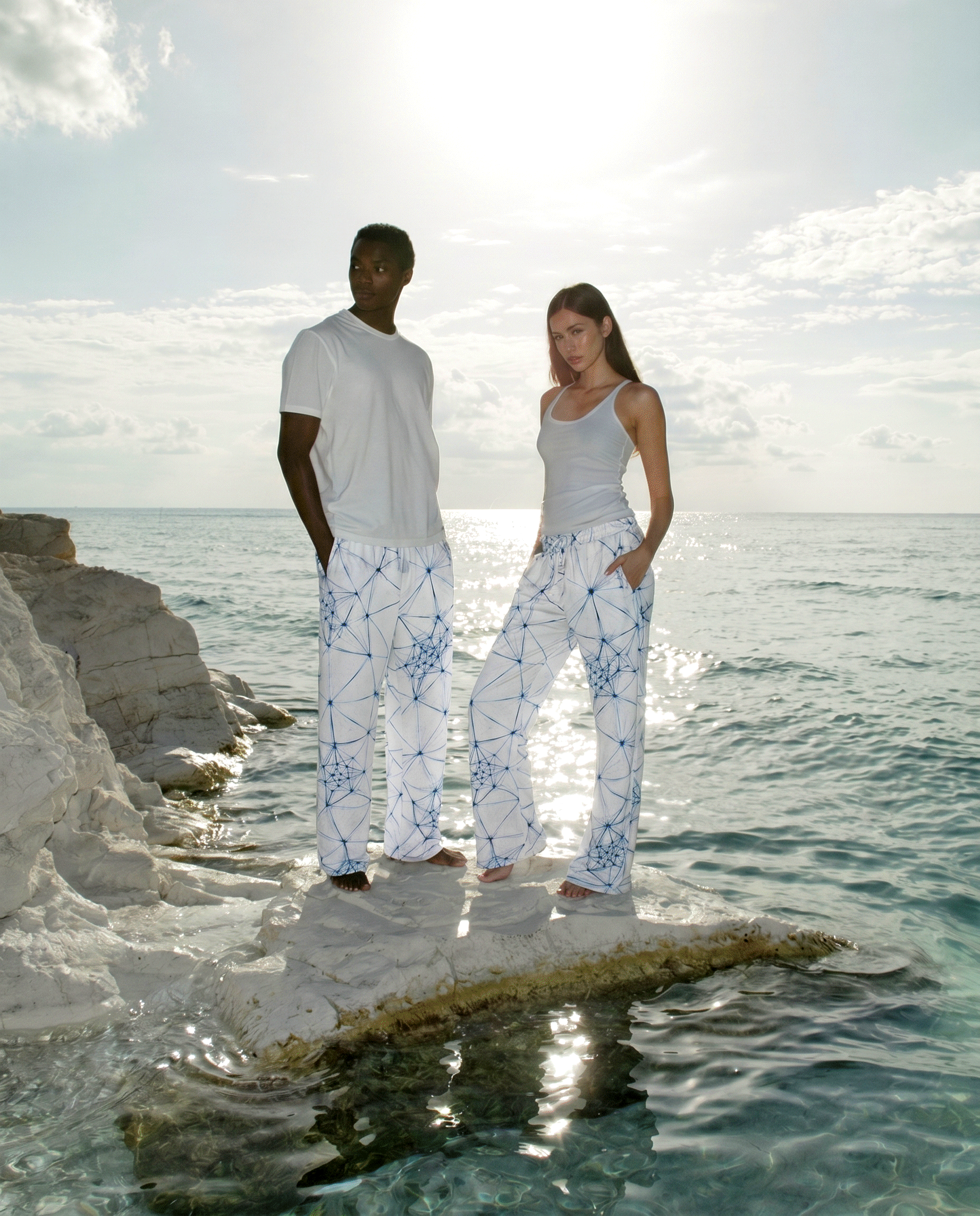 Two people standing on a rock by the ocean wearing white ANSO joggers with blue patterns.