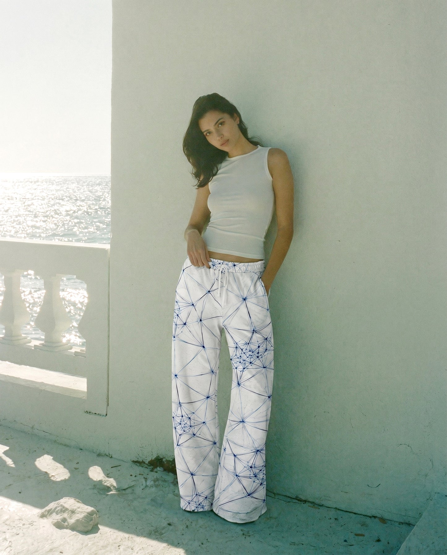 Woman wearing a white top and patterned pants leaning against a white wall by a window with a view of water.