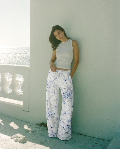 Woman wearing a white top and patterned pants leaning against a white wall by a window with a view of water.